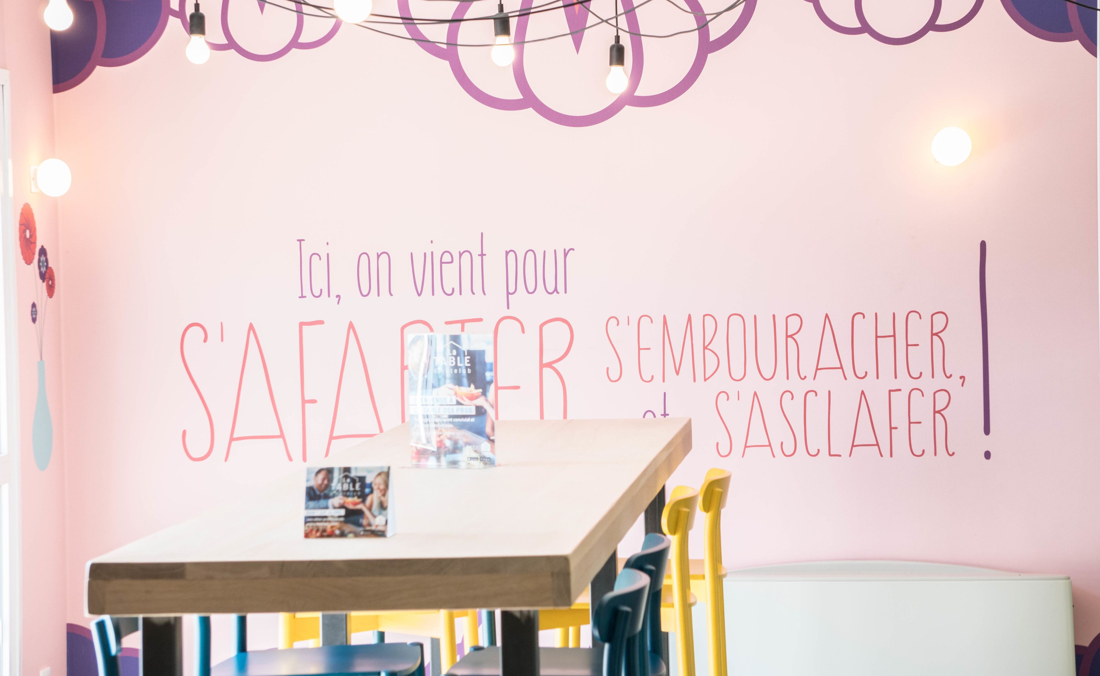 A room with a pink wall, a wooden table, and yellow chairs. There is a poster on the wall with text in French.