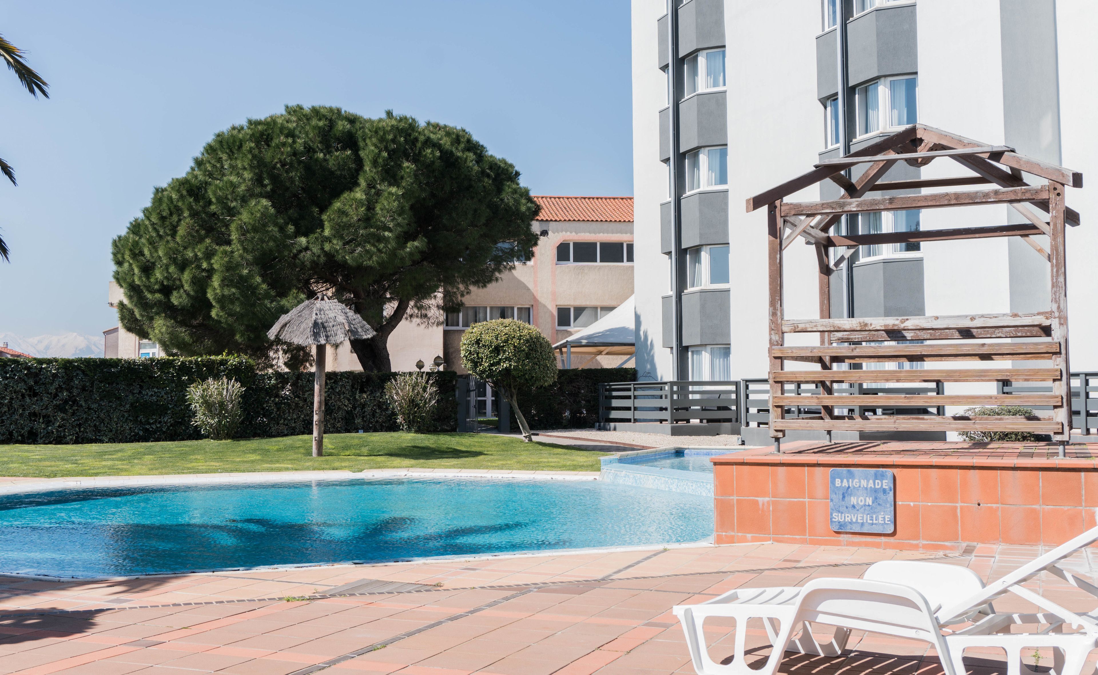 Una piscina con una pérgola de madera y sillones de descanso, rodeada de un césped cuidado y un árbol grande.