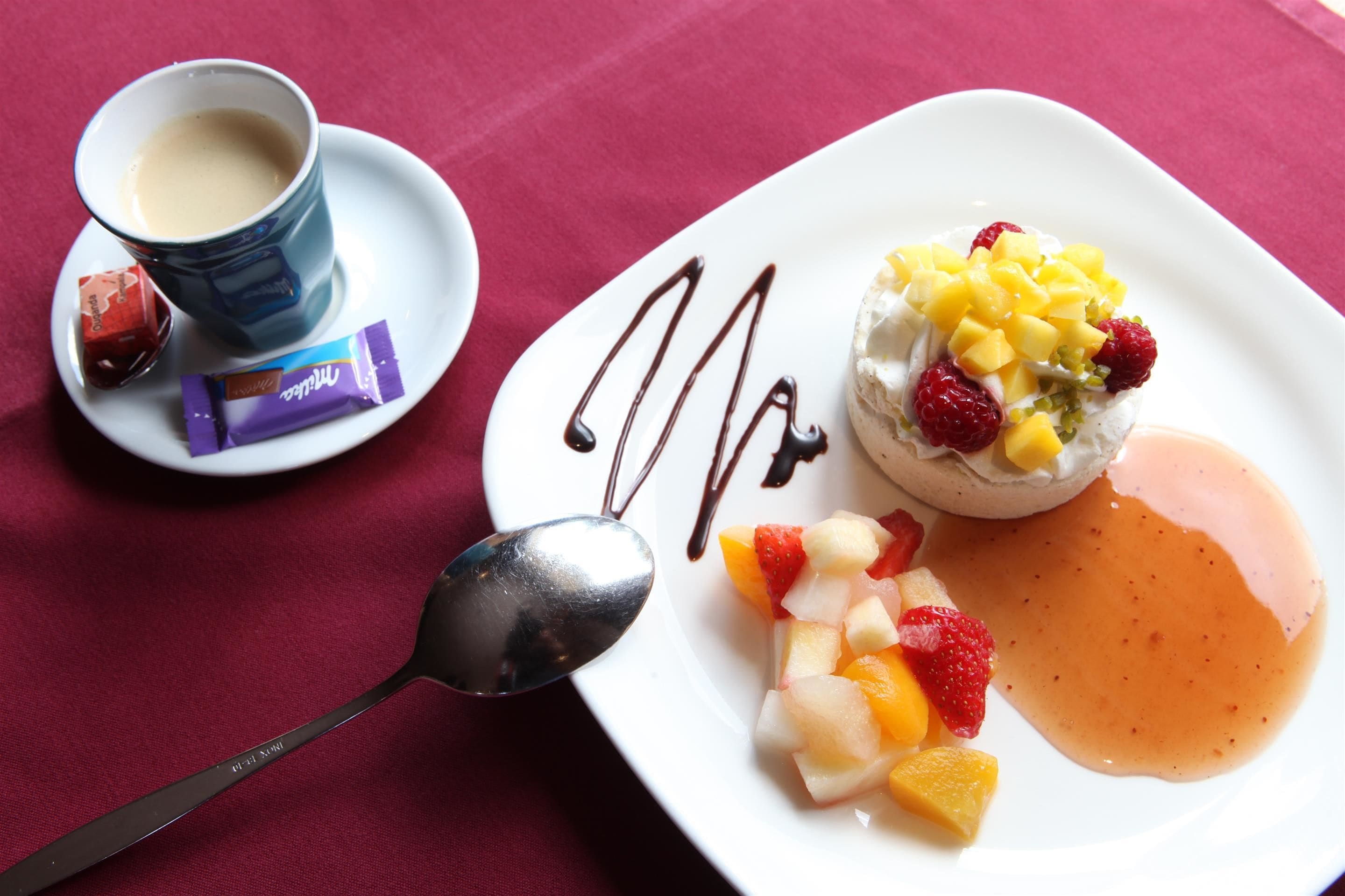 A dessert plate with fruit and chocolate sauce, a cup of coffee, and a spoon on a red tablecloth.