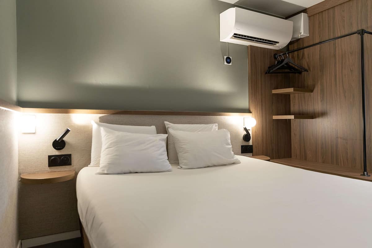 A neatly arranged hotel room with a bed, bedside tables, a wall-mounted air conditioner, and a coat rack.