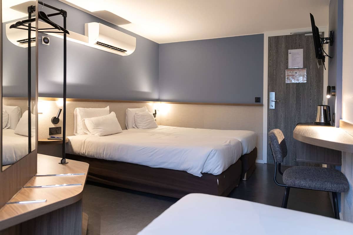 A modern hotel room with a large bed, bedside tables, a mirror, a desk, a chair, a small table, a wall-mounted TV, and a built-in sink.