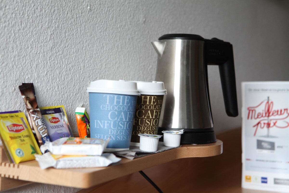 A wooden shelf with a coffee maker, two insulated mugs, a box of Lipton tea, a packet of instant coffee, and a small container of milk.