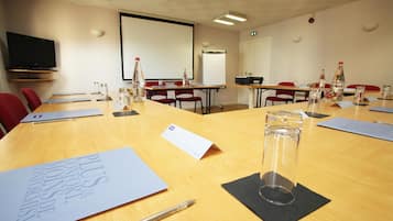 A conference room with a long table, chairs, a projector screen, a television, and various documents and water bottles.