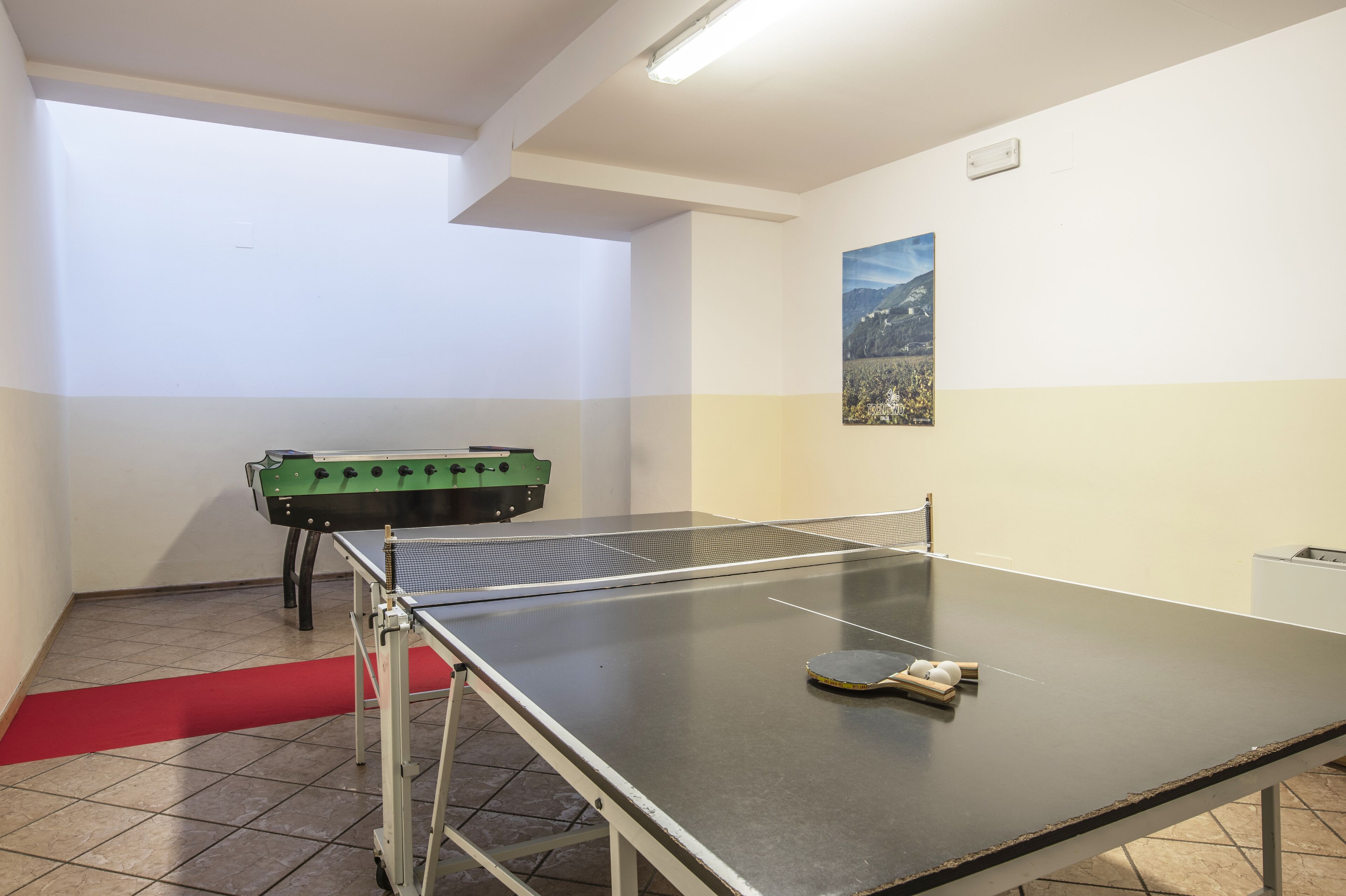 A ping pong table with a net, a foosball table, and a red carpet in a room with tiled flooring and a single wall-mounted picture.