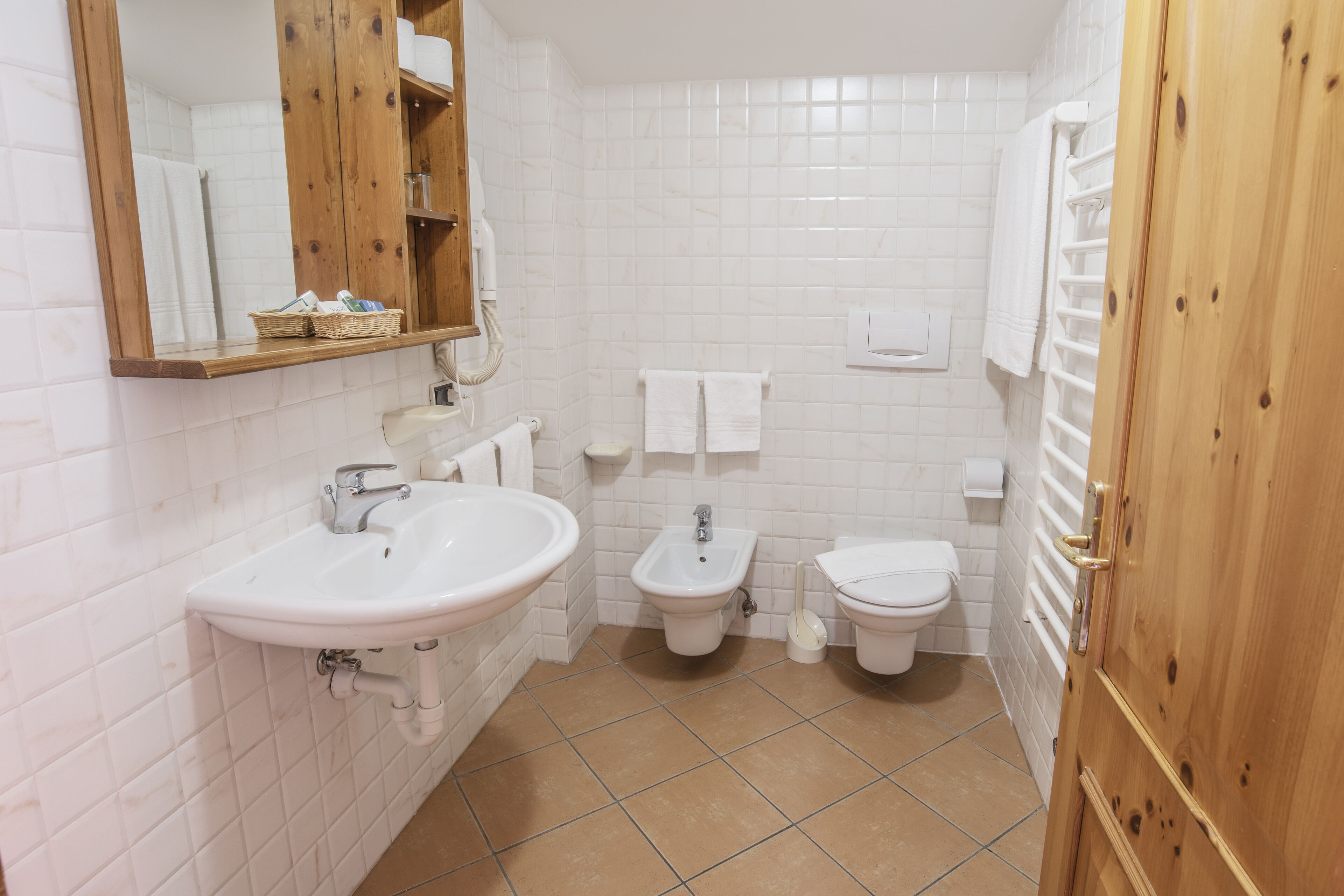 A bathroom with a sink, bidet, and toilet, white tiled walls, and a wooden door.