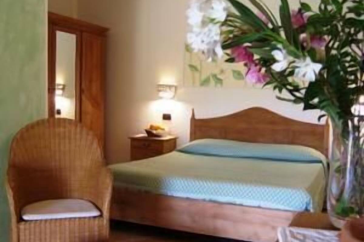 A bedroom with a bed, a nightstand, a wicker chair, and a vase with flowers.