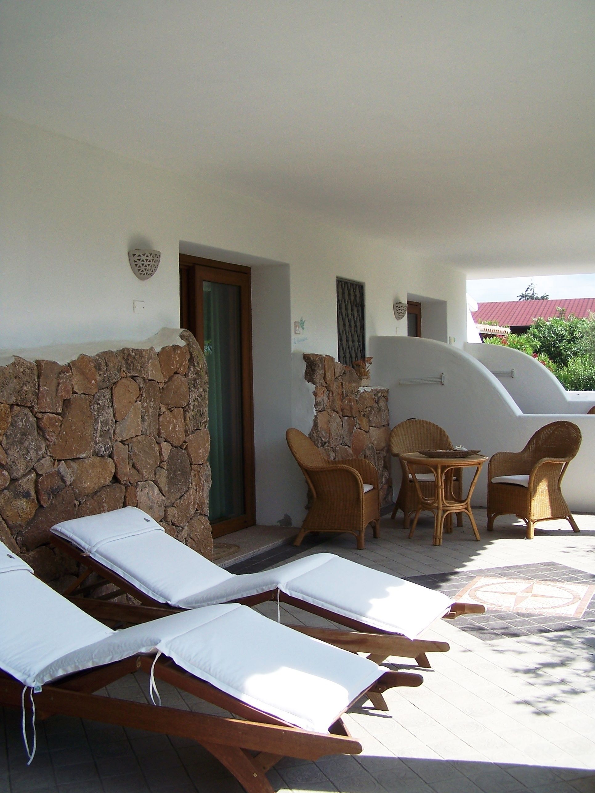 Un'area esterna luminosa con sedute relax e un tavolo, pareti in pietra e una porta che dà su un ambiente interno.