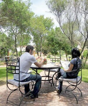 Two people sitting at a round table outdoors with metal chairs, surrounded by trees. - Club Hotel Ragno D'Oro (Aglientu)