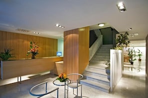 A modern reception area with a wooden reception desk, a staircase, and a seating area with a vase of flowers. - Nautico (Santa Cruz de Tenerife)
