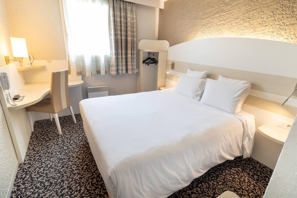 A hotel room with a large bed, a desk with a chair, a bedside table, a hanging closet, and a window with curtains.