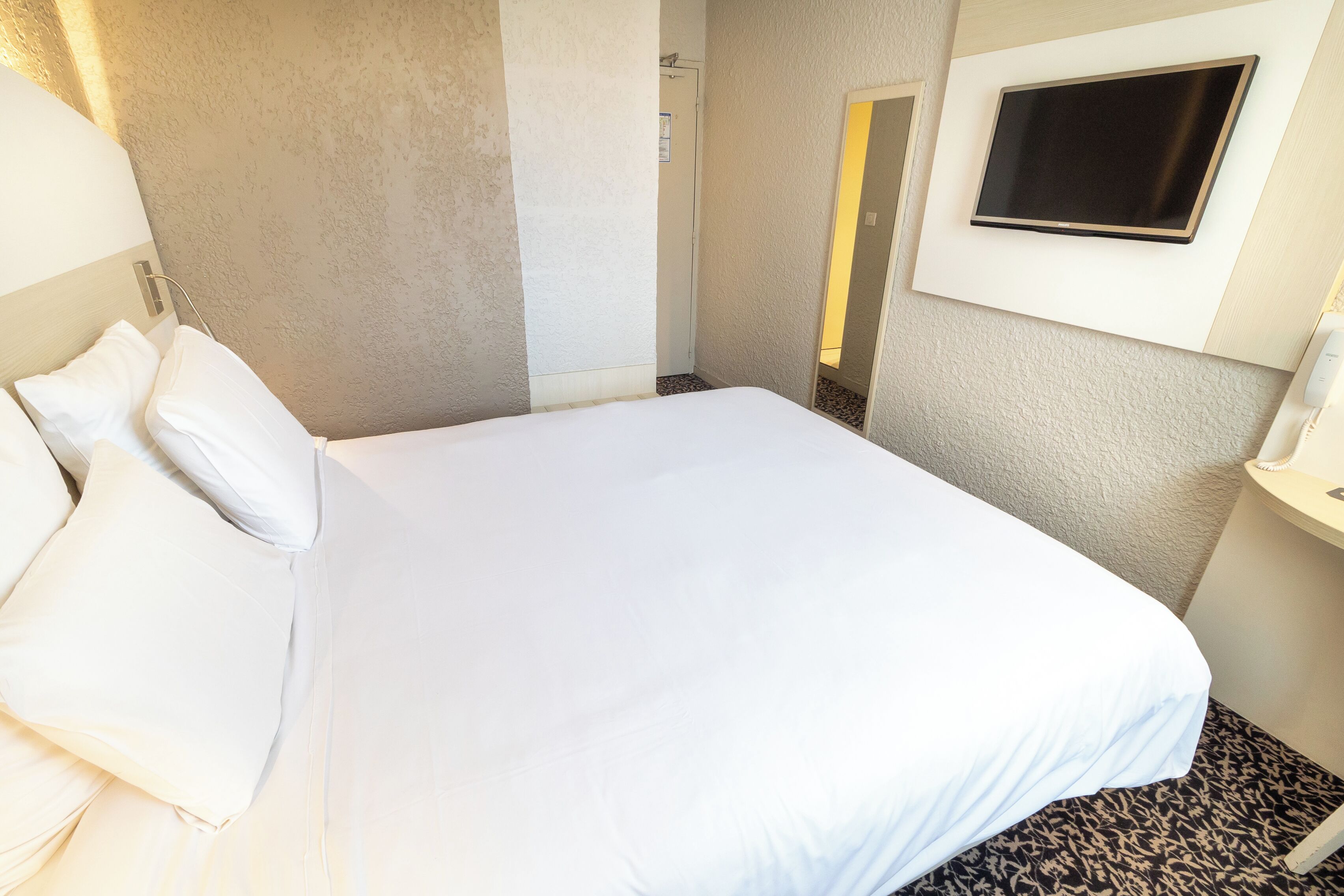 A hotel room with a bed, a TV mounted on the wall, and a bathroom visible through an open door.