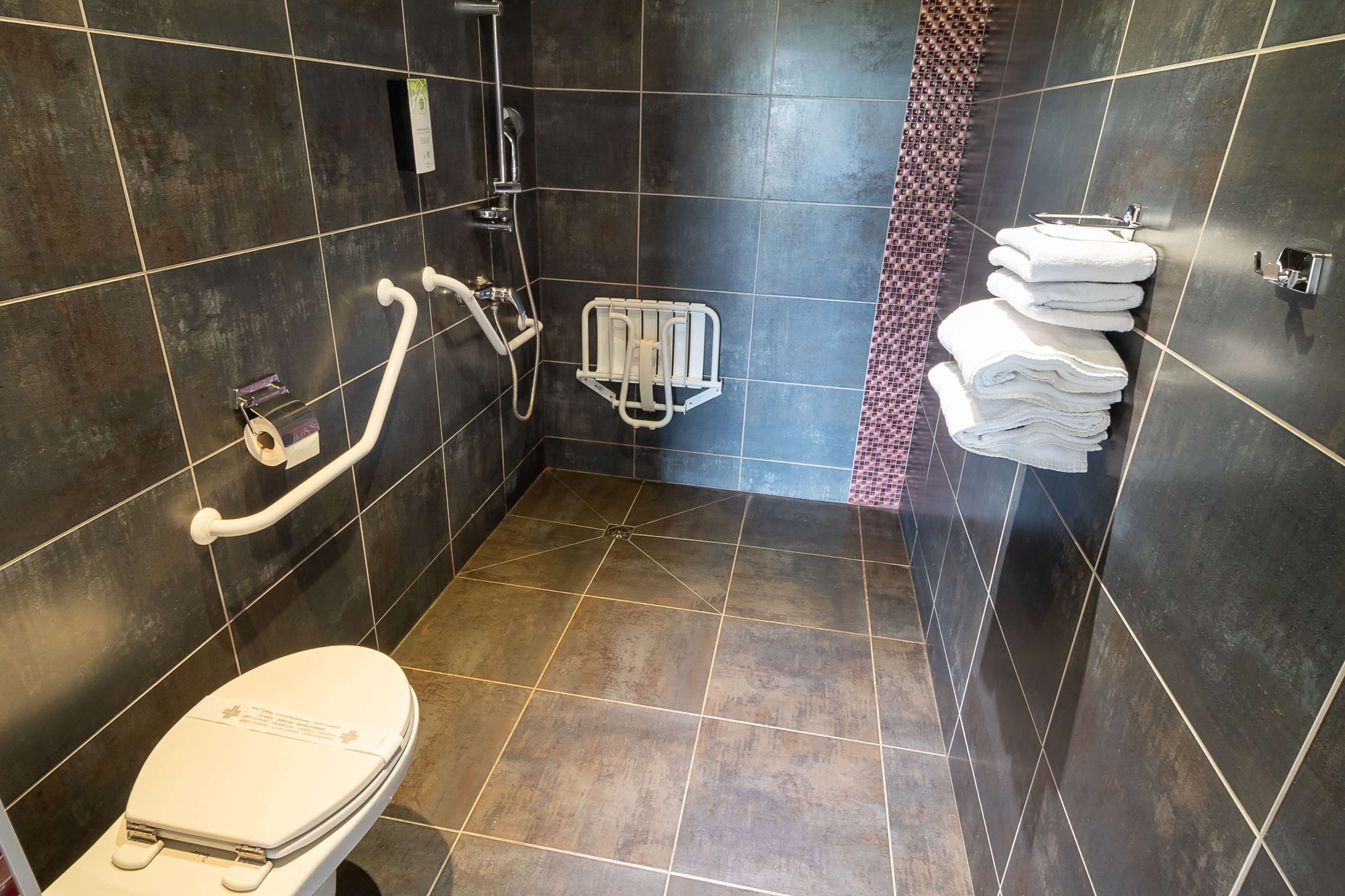 A bathroom with a toilet, a shower with a handrail, a towel rack, and a tiled floor.