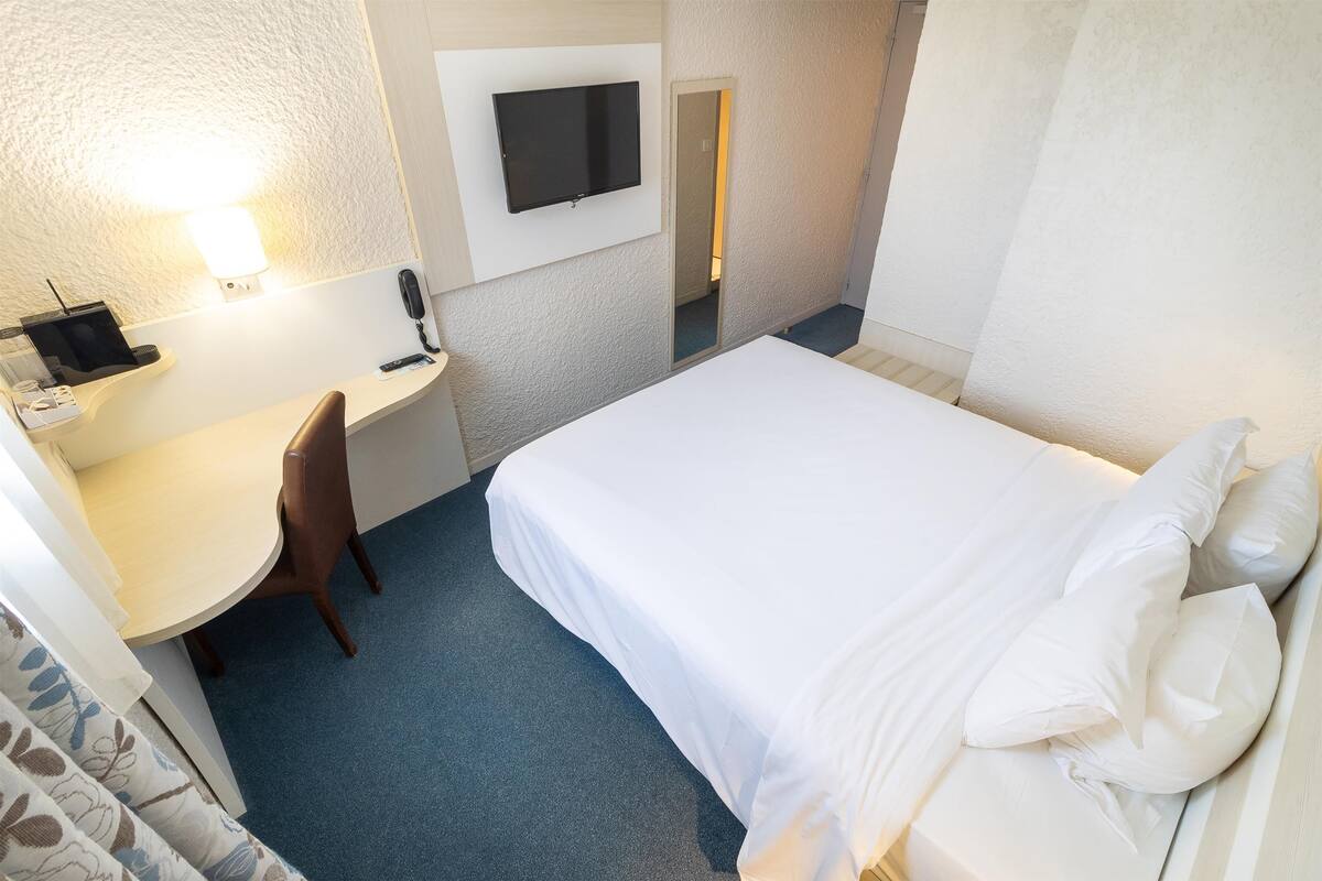 A hotel room with a bed, a desk, a chair, a TV mounted on the wall, and a bathroom visible through a door.