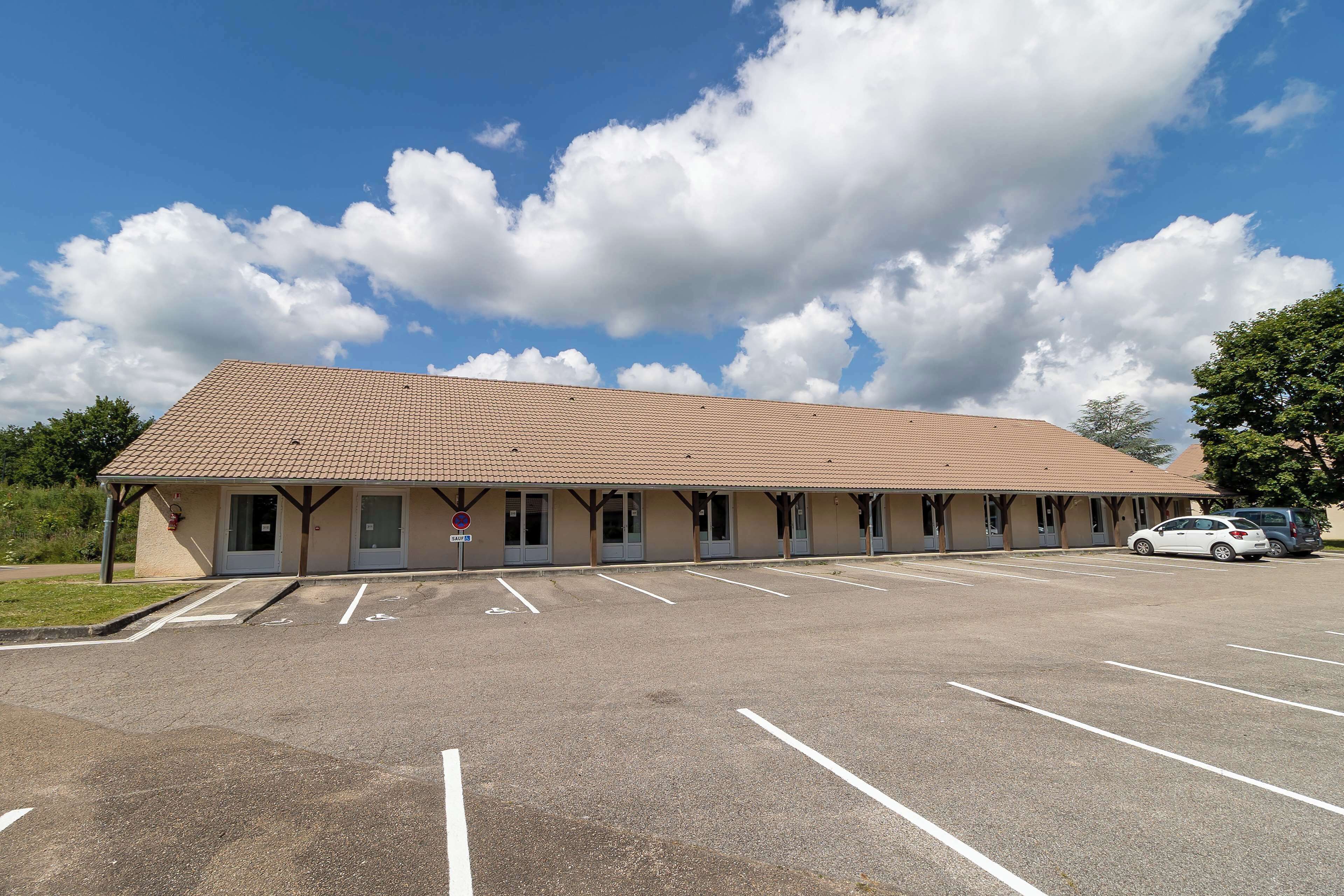 A building with a tiled roof and multiple doors, a parking lot with marked spaces, and a clear blue sky with clouds.