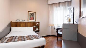 Una habitació d'hotel amb llit, escriptori, cadira, planta en test i un quadre a la paret.