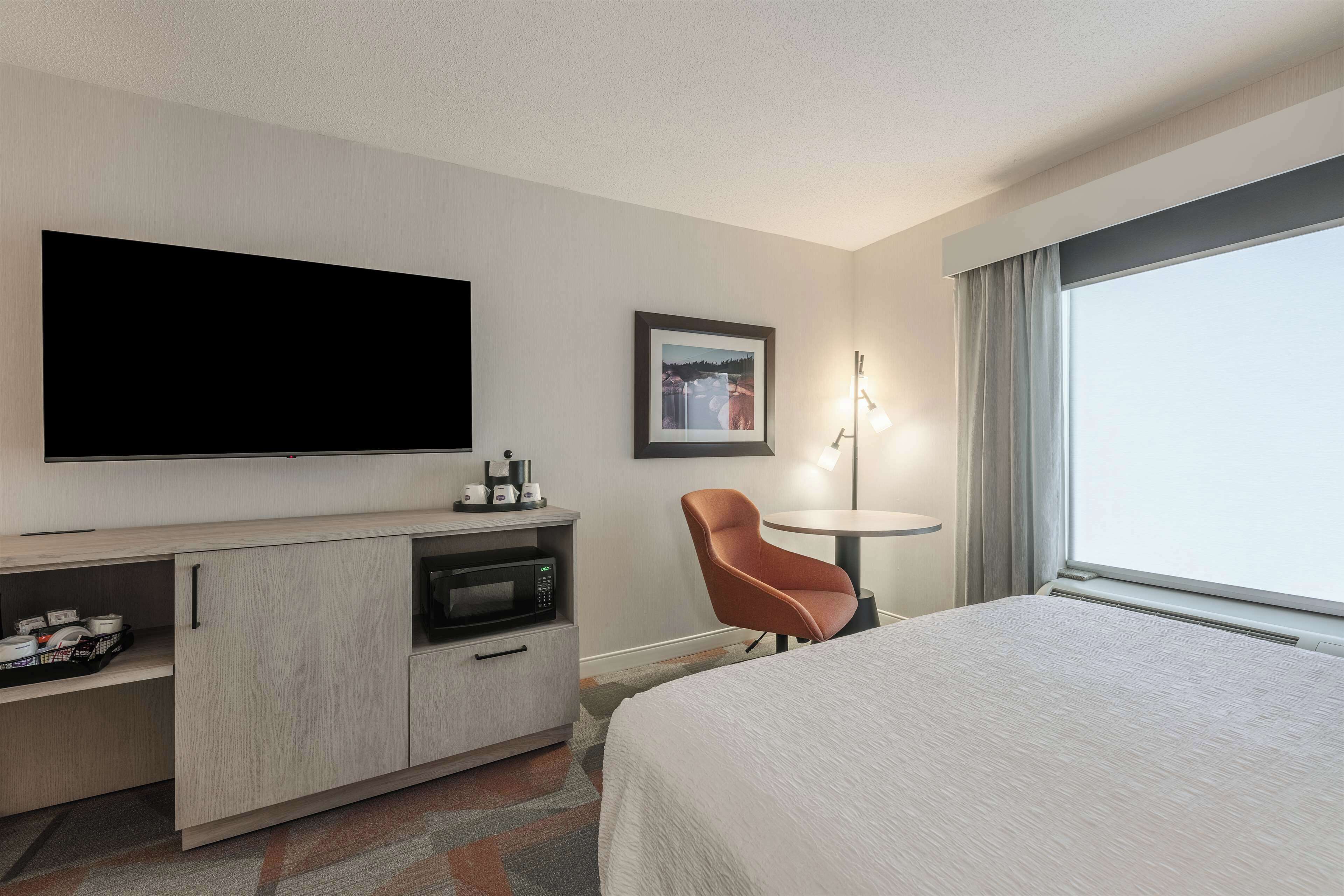 Habitación de hotel con cama, televisión, microondas, una mesita con una silla y una ventana con cortinas.