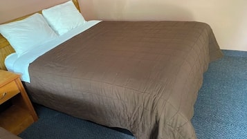 Una cama individual con una colcha marrón y almohadas blancas, una mesita de noche de madera y un suelo cubierto de moqueta azul.