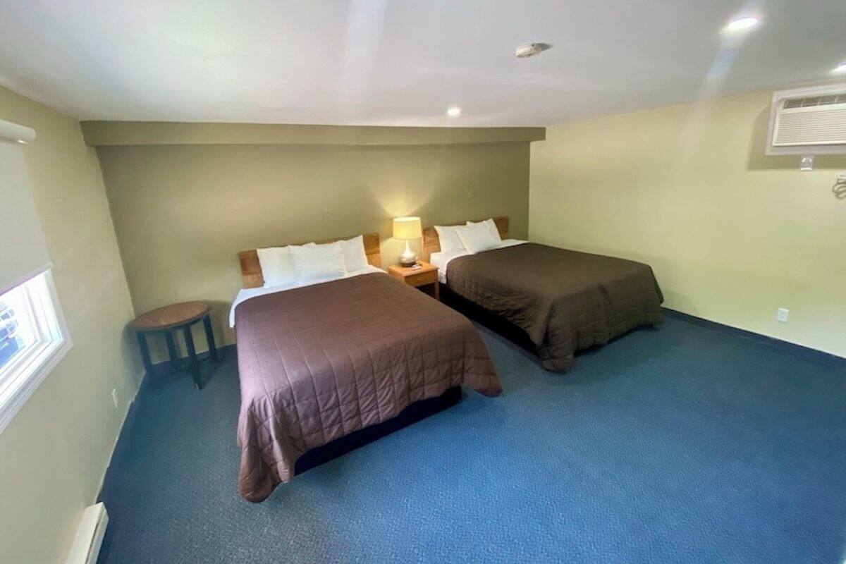 A room with two beds, a small stool, a nightstand with a lamp, and a window.