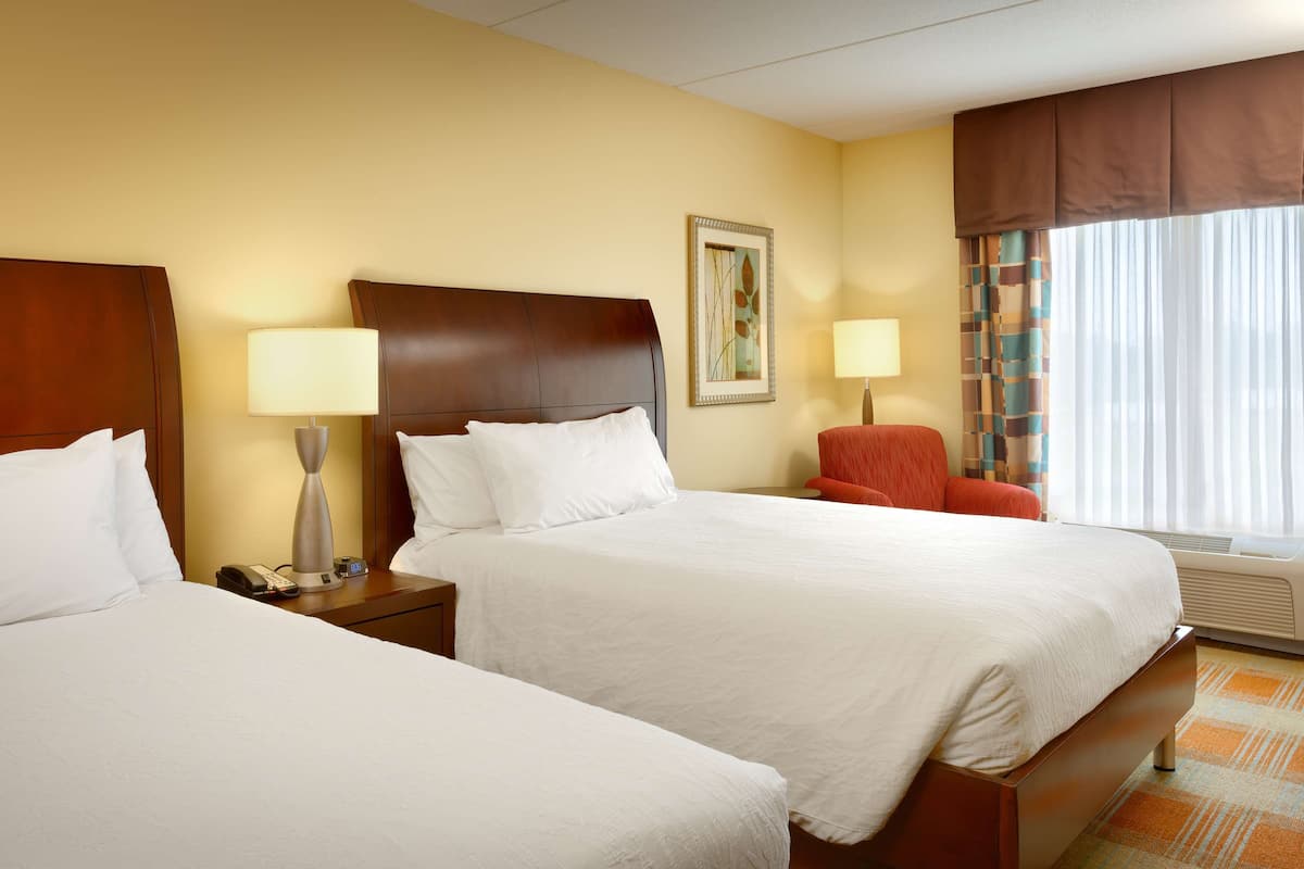 A hotel room with two beds, a nightstand with a lamp, a chair, a painting on the wall, and a window with curtains.