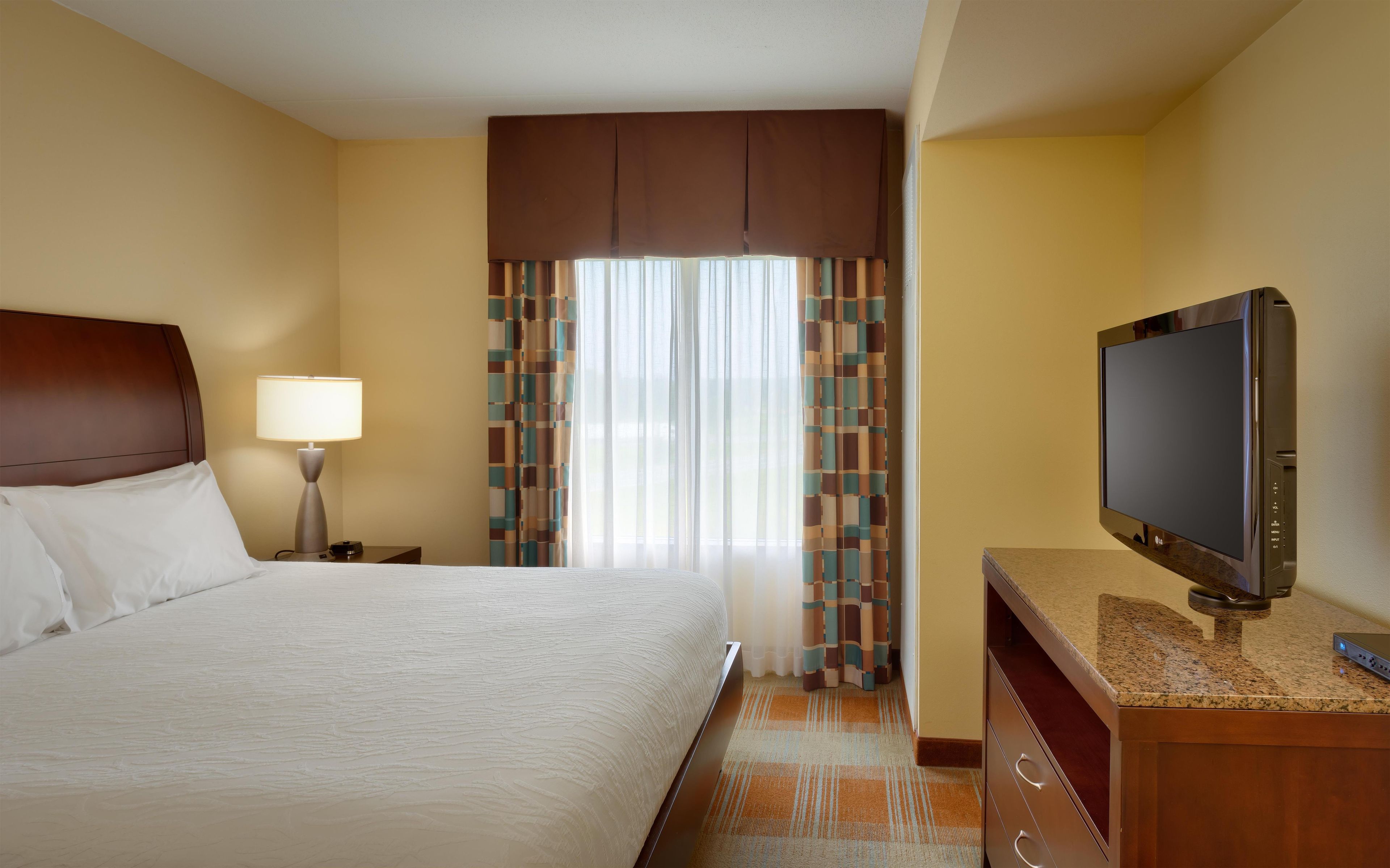Une chambre d’hôtel avec un lit, une télévision, une table de chevet, une lampe et une fenêtre avec des rideaux.