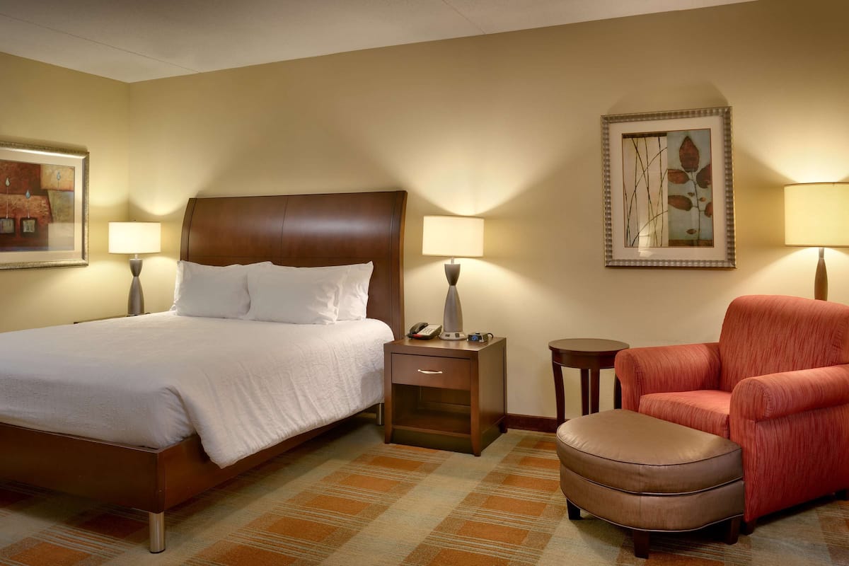 A hotel room with a bed, bedside tables, a chair, an ottoman, and artwork on the walls.