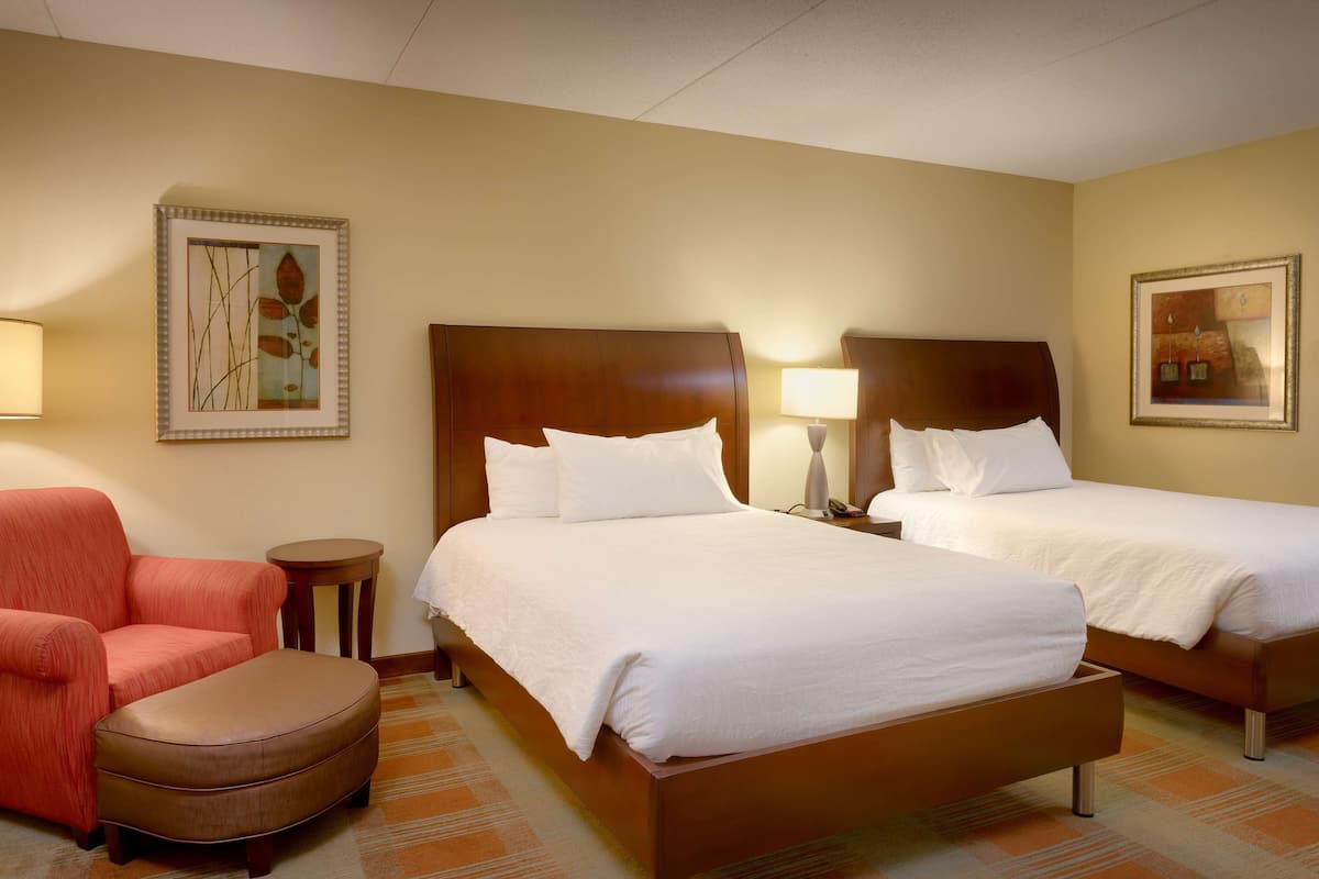 A hotel room with two beds, a red armchair, a small round table, and two wall-mounted lamps.