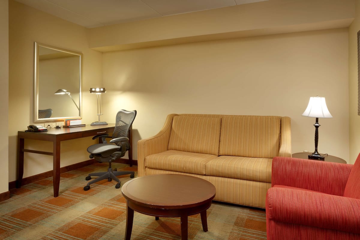 A hotel room with a sofa, a red armchair, a desk with a lamp, a chair, and a round table.