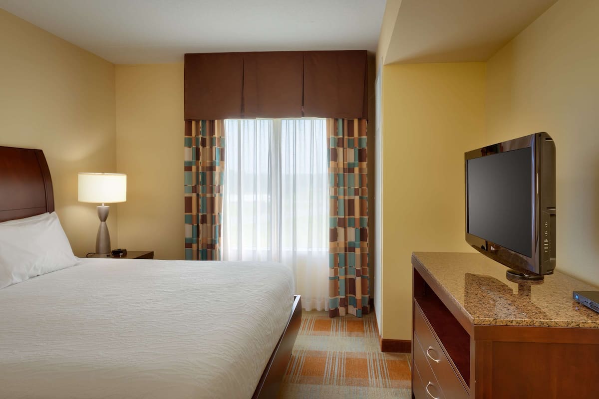 A hotel room with a bed, a TV, a nightstand, a lamp, curtains, and a window.