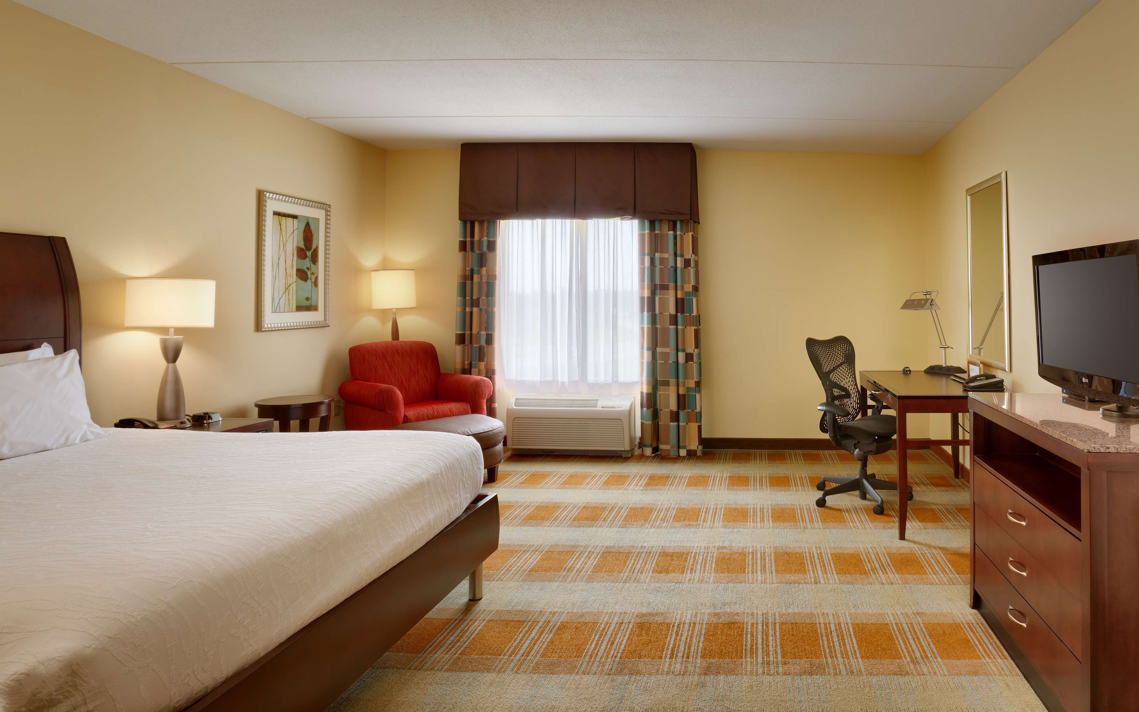 A hotel room with a bed, a desk with a computer, a chair, a television, and a window with curtains.