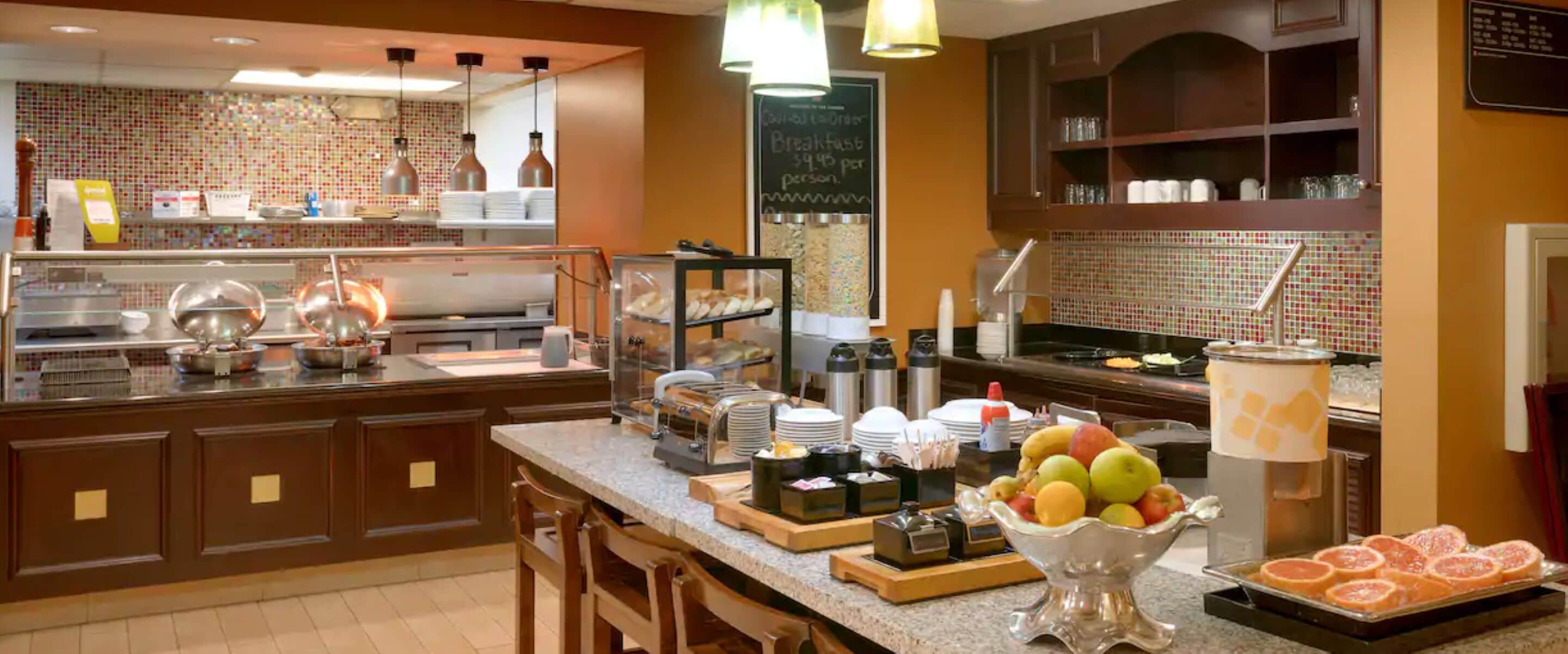A buffet breakfast area with a variety of food items, including fruits and pastries, and a chalkboard menu indicating breakfast prices.