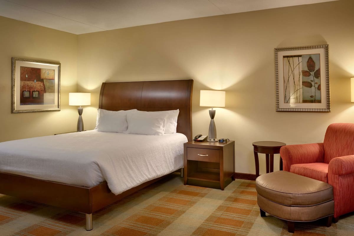 A hotel room with a bed, bedside table, lamp, chair, ottoman, and artwork on the walls.