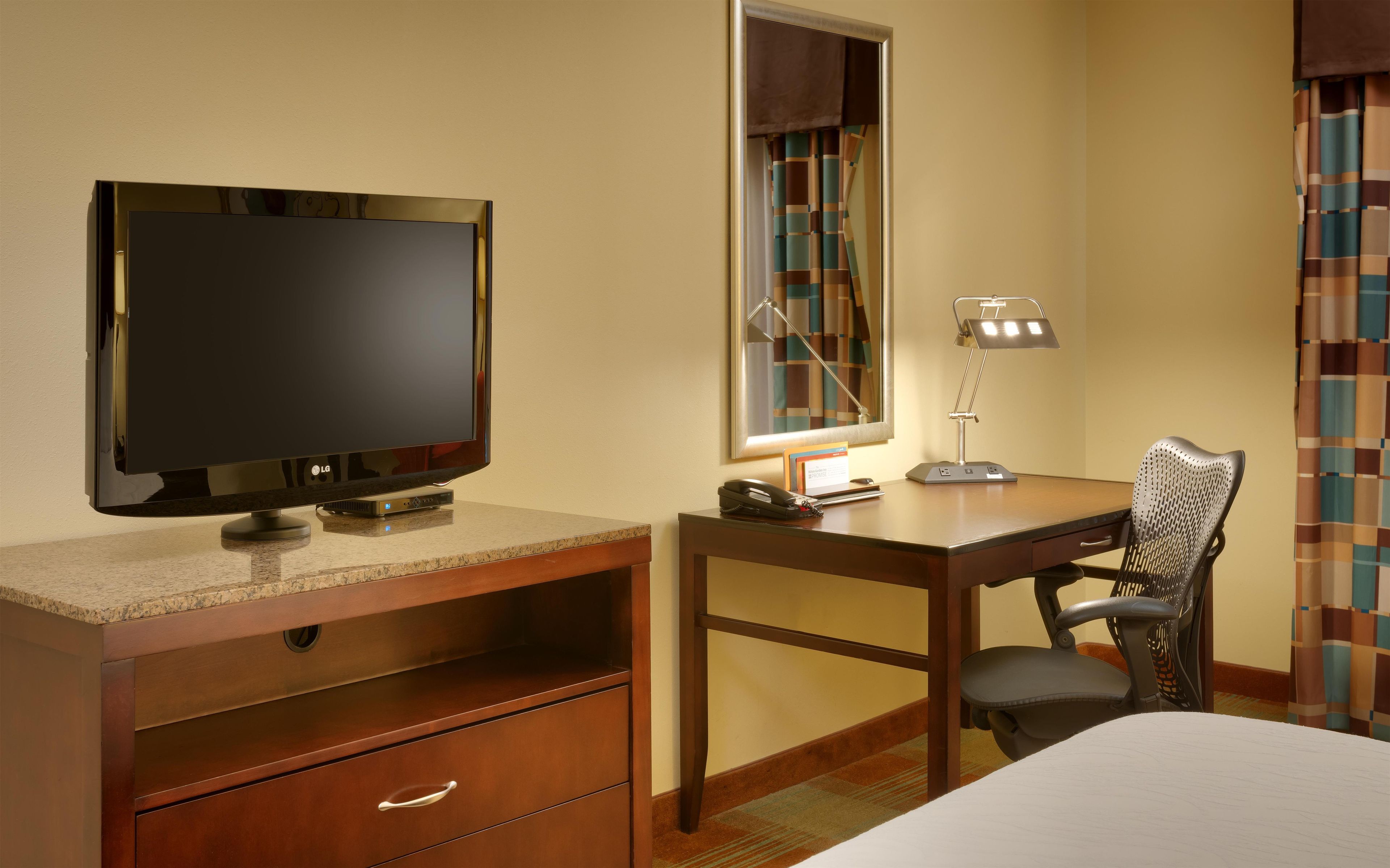 A hotel room with a flat-screen TV, a desk with a lamp, a chair, and a bed.