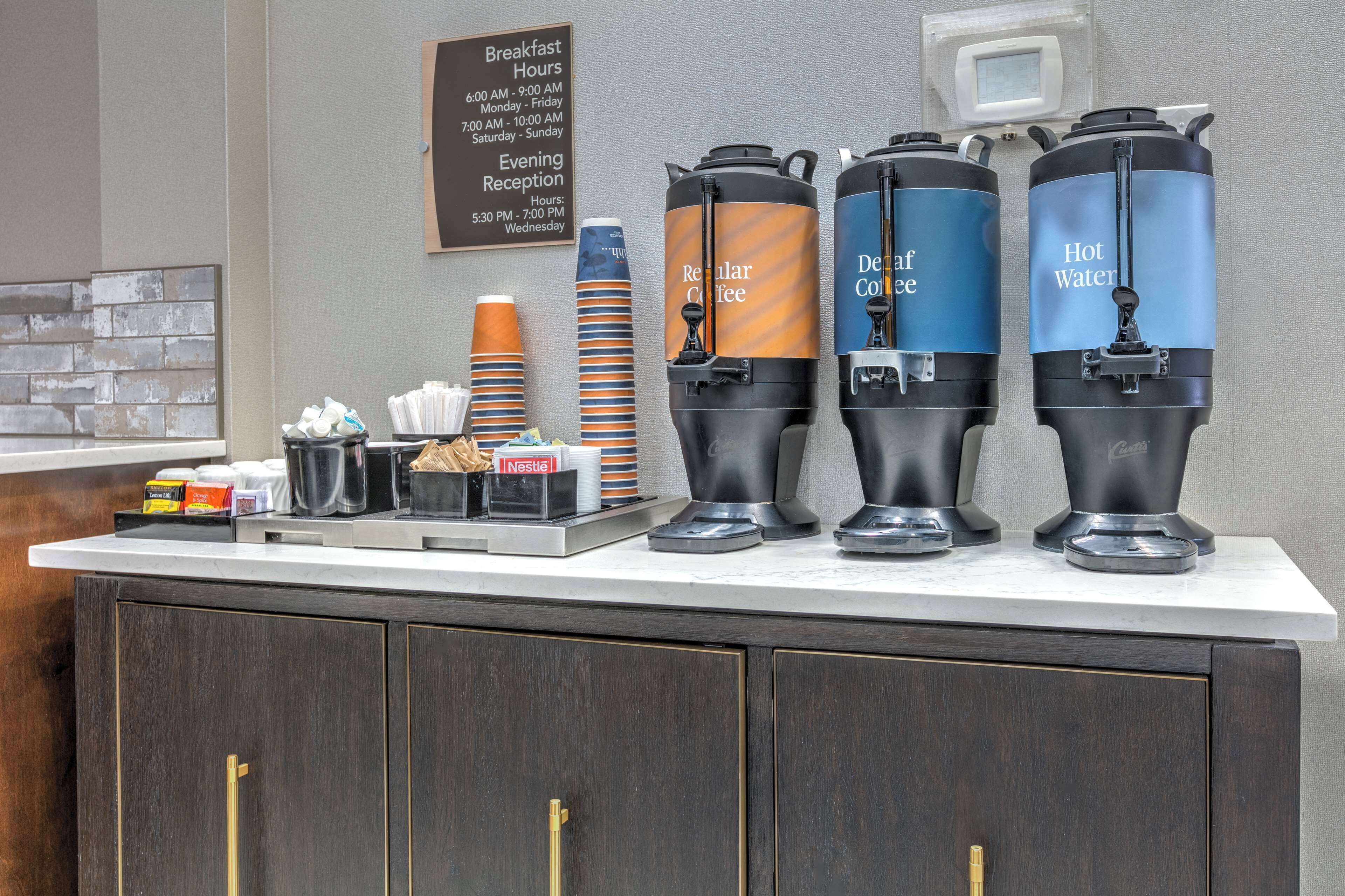 Un comptoir pour le petit-déjeuner avec des distributeurs de café et d’eau, un panneau indiquant les heures du petit-déjeuner, ainsi que divers articles pour le petit-déjeuner.