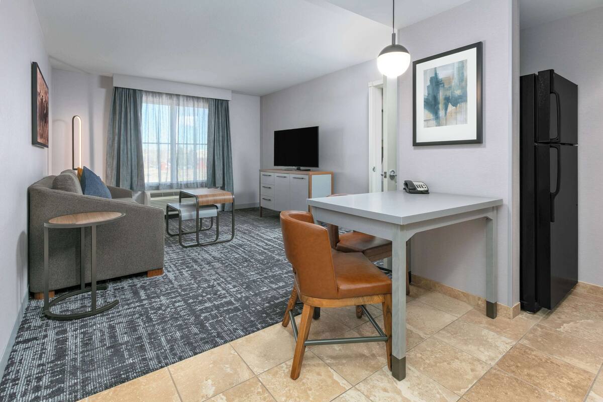 Une chambre d’hôtel moderne avec un canapé gris, une petite table ronde, une chaise en bois, un bureau avec une chaise, un téléviseur à écran plat, un réfrigérateur et un tableau encadré au mur.