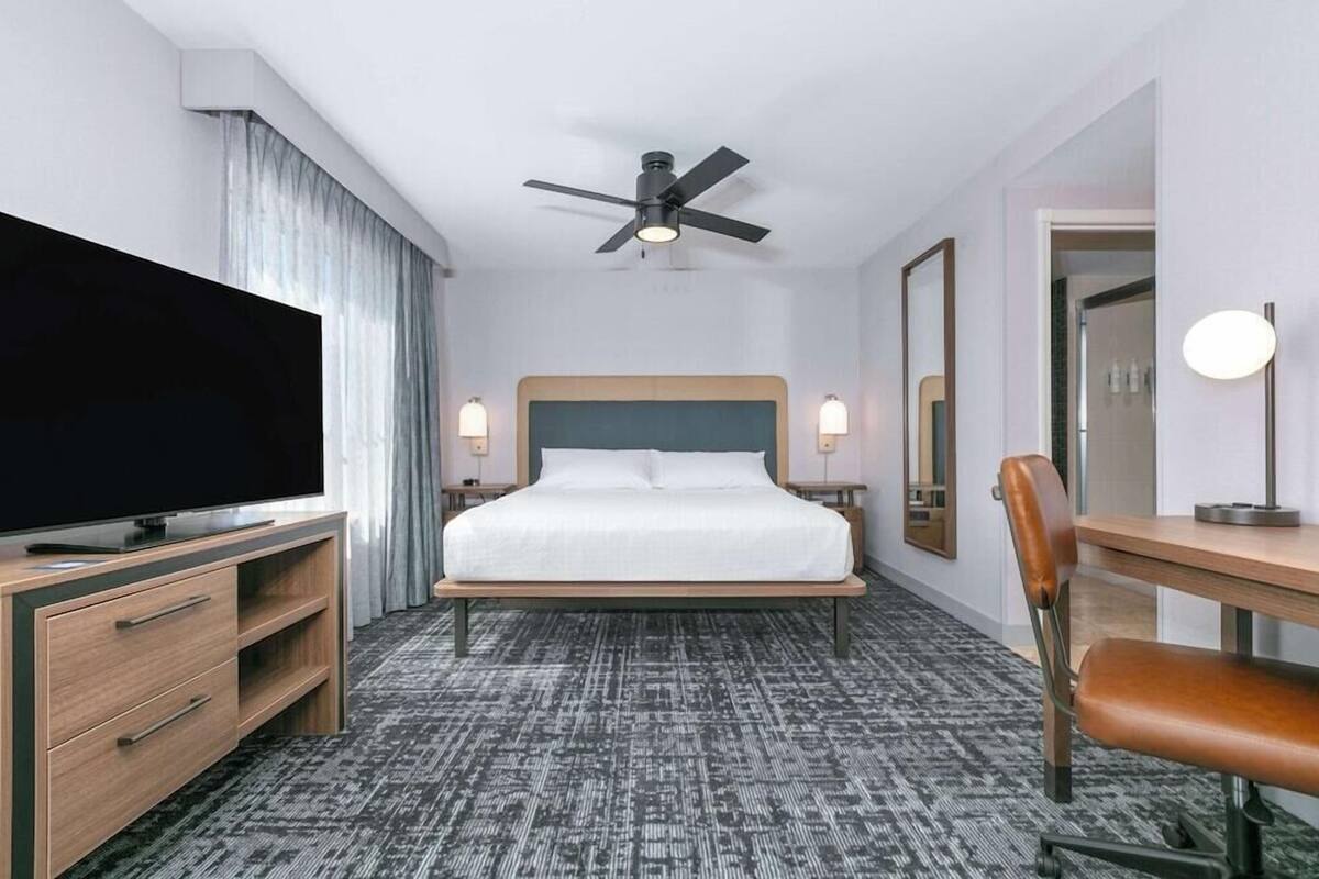 Een hotelkamer met een bed, een televisie, een bureau, een stoel, een plafondventilator en een spiegel.