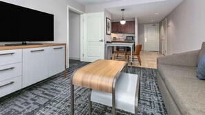 Un salon moderne avec un téléviseur à écran plat, une table basse en bois, un canapé gris et une cuisine en arrière-plan.