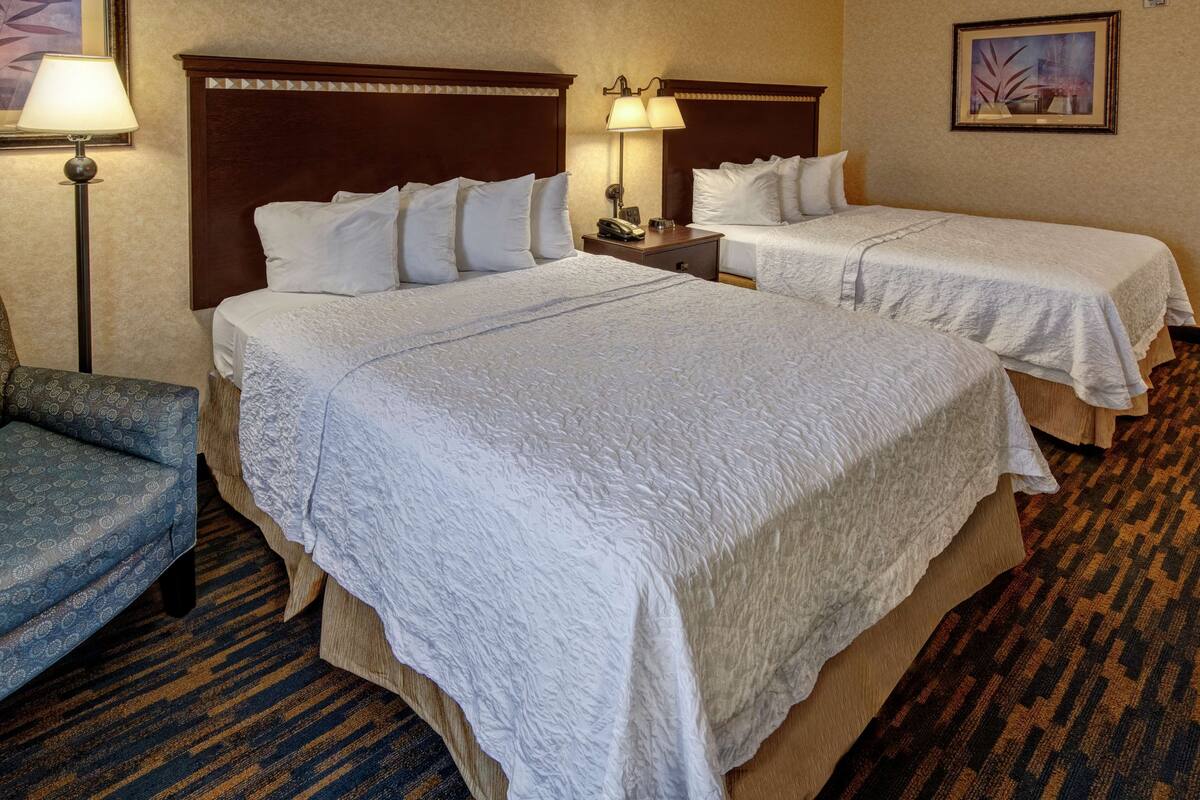 Quarto de hotel com duas camas, uma mesa de cabeceira, um abajur, uma cadeira e piso coberto por carpete.