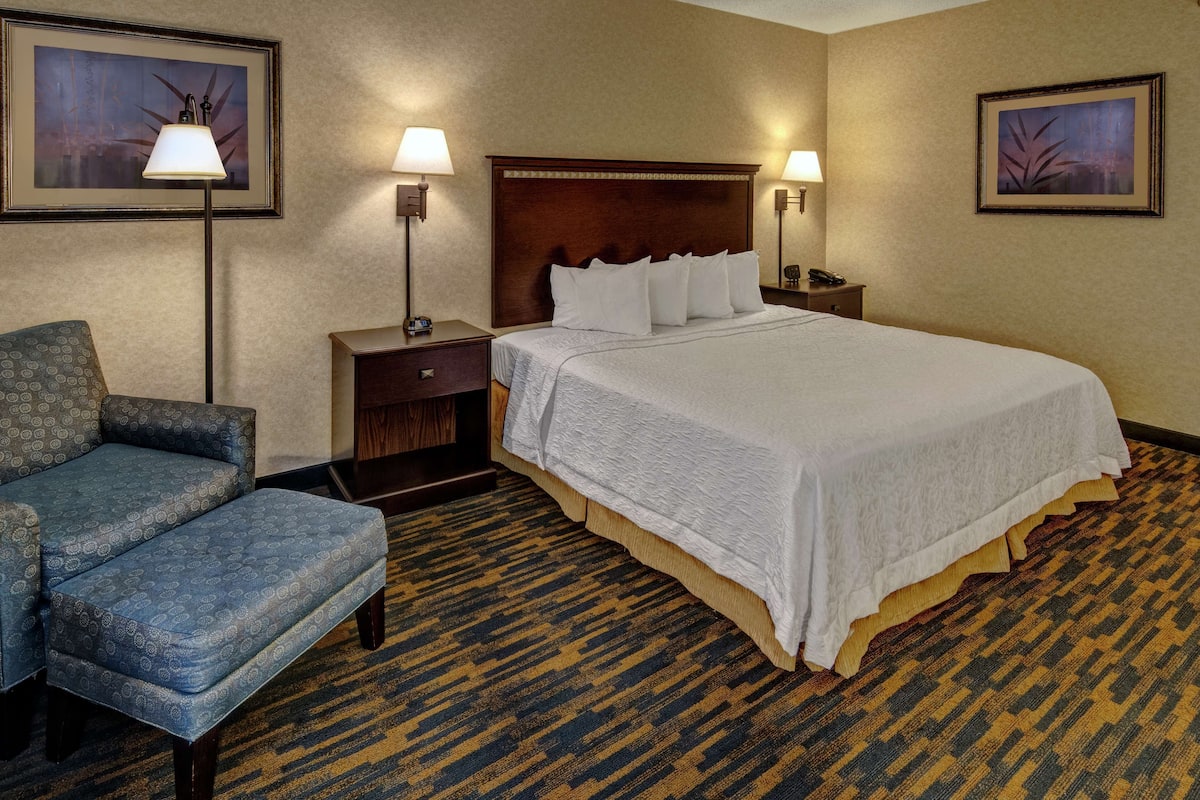 Quarto de hotel com uma cama grande, uma cadeira, duas mesas de cabeceira com luminárias e duas quadros nas paredes.