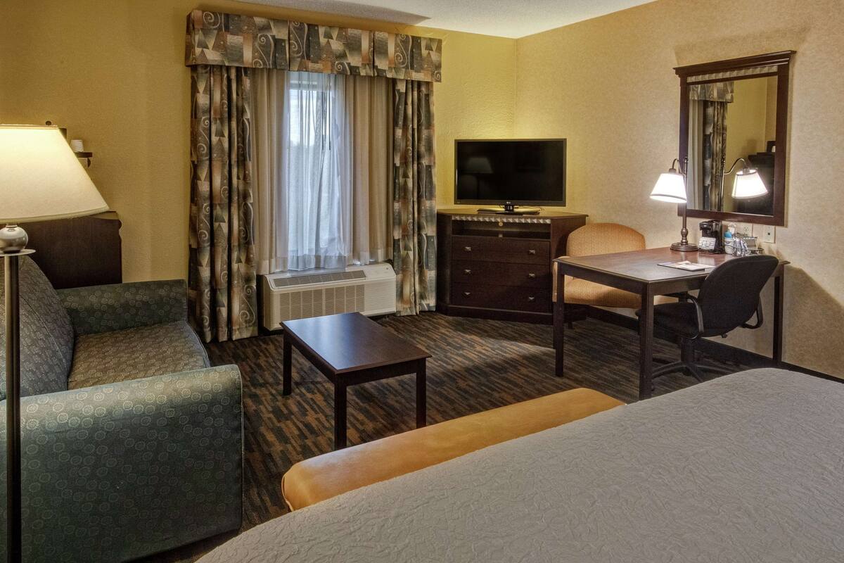 Quarto de hotel com cama, escrivaninha, cadeira, televisão, luminária, penteadeira e janela com cortinas.