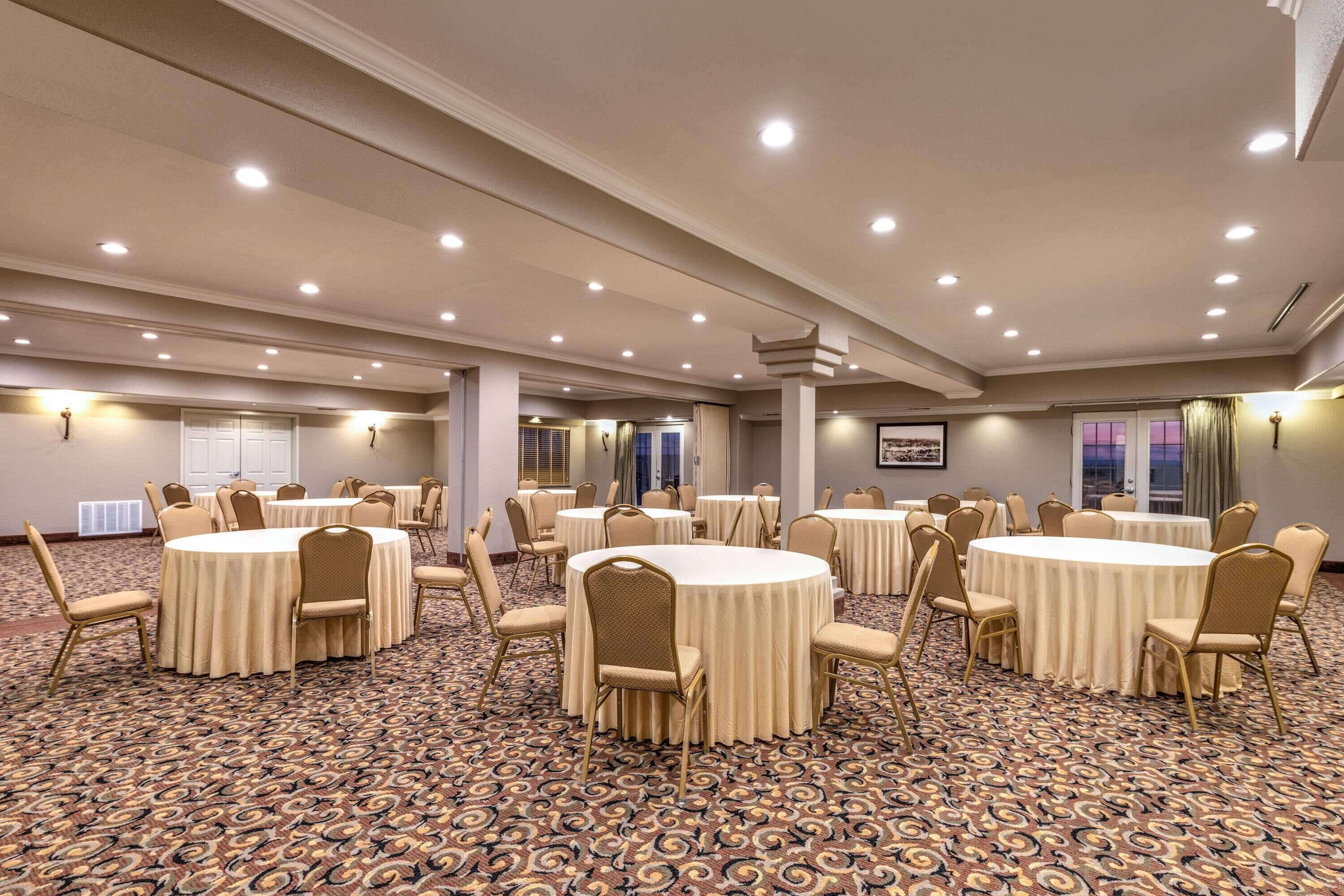 Un salón de conferencias amplio con varias mesas redondas, sillas color beige y una alfombra estampada.