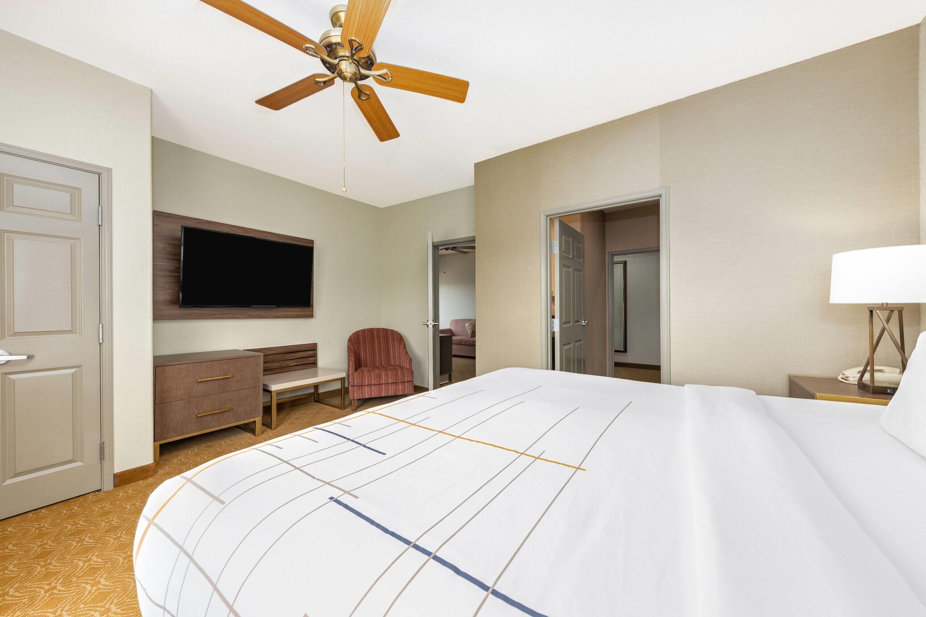 A hotel room with a large bed, a ceiling fan, a TV mounted on the wall, a desk with a lamp, and a chair.