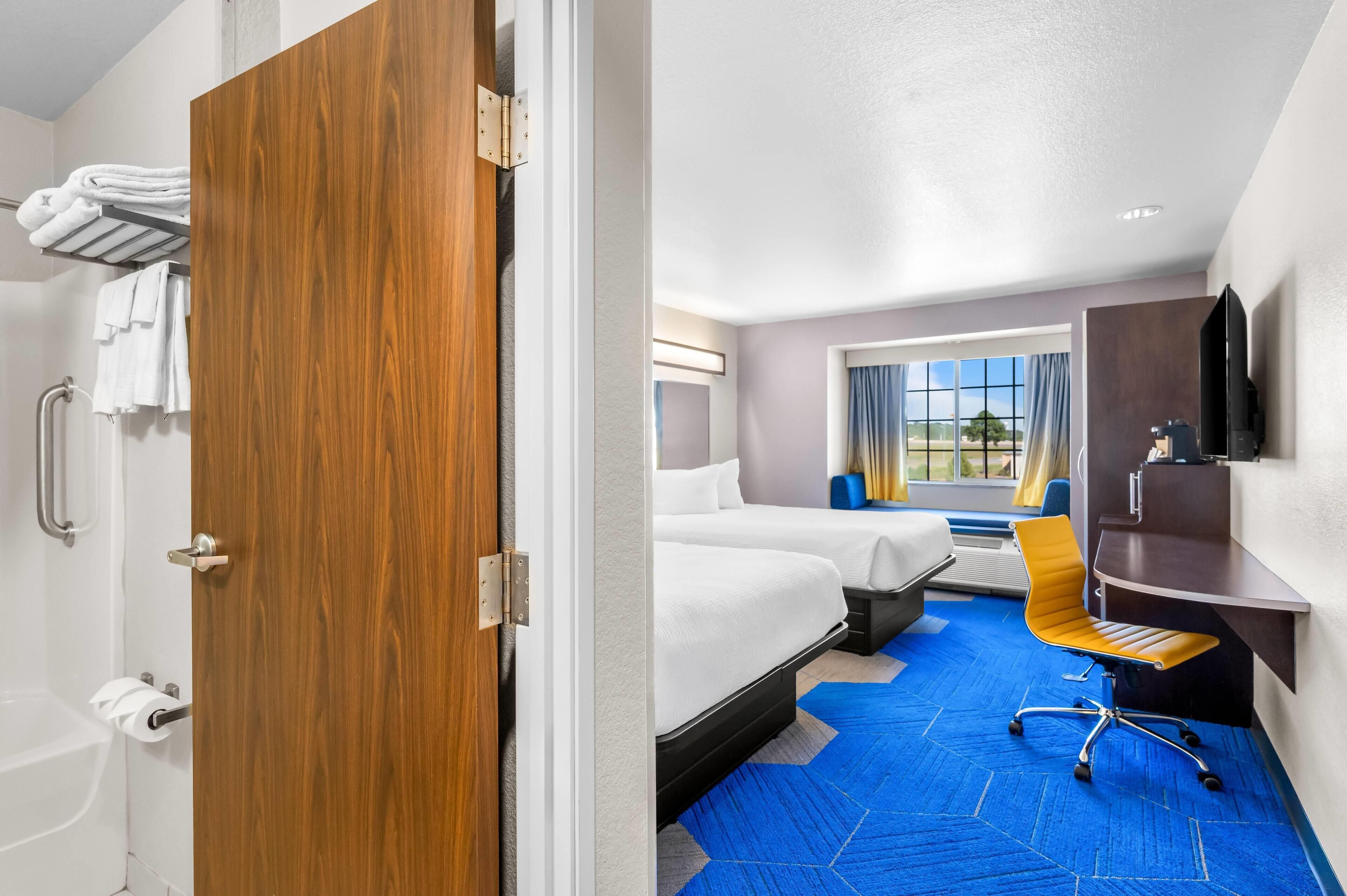 Une chambre d’hôtel avec une porte en bois, un lit, un bureau, une chaise et une grande fenêtre donnant sur un beau paysage.
