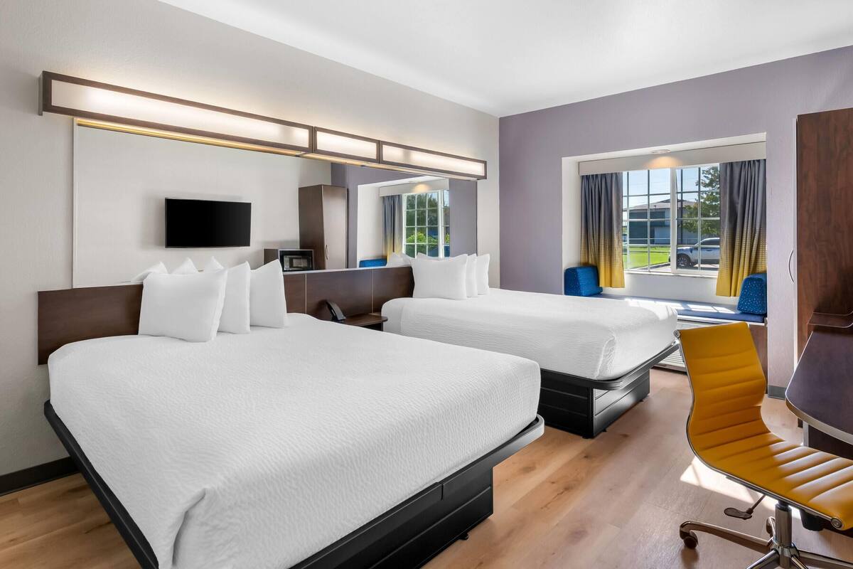 Ein Hotelzimmer mit zwei Betten, einem Schreibtisch, einem Stuhl, einem Fernseher und einem Fenster mit Vorhängen.