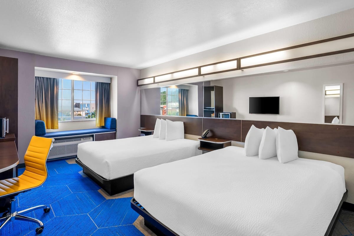 Ein Hotelzimmer mit zwei Betten, einem Schreibtisch, einem Stuhl und einem großen Fenster mit Vorhängen.