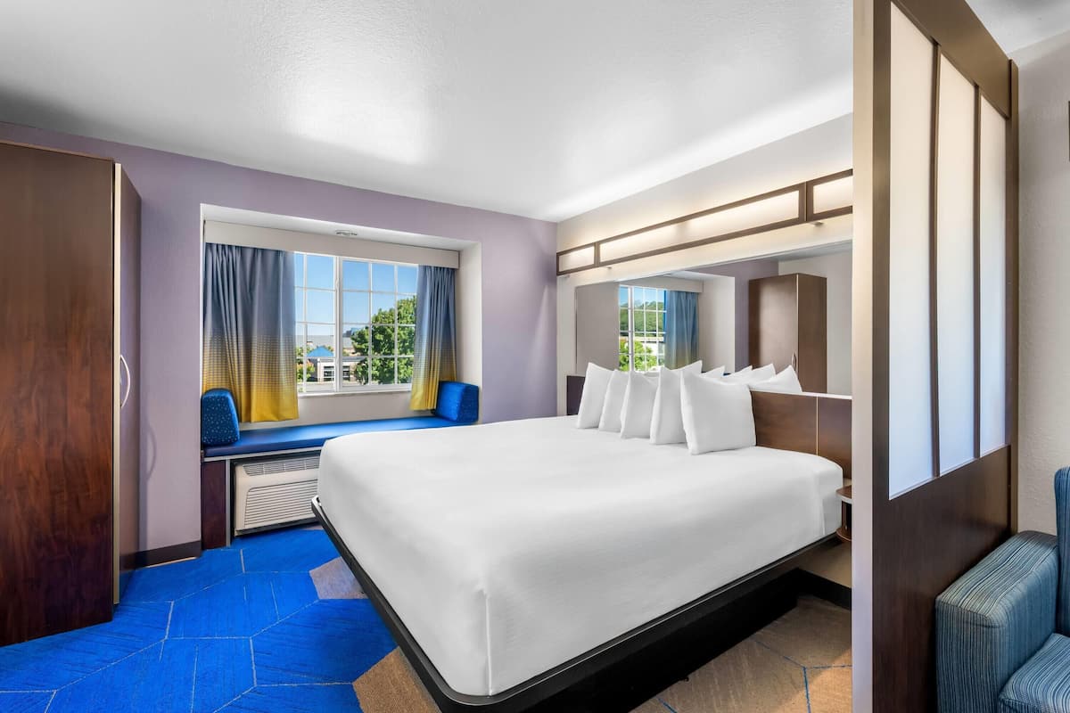 Ein Hotelzimmer mit einem großen Bett, einem Fenster mit Vorhängen, einer Sitzecke mit einem Sessel und einem Spiegel.