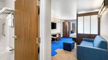 Une chambre d’hôtel avec une porte en bois, une salle de bain avec une baignoire, un bureau avec une chaise, un canapé bleu et une grande fenêtre avec des rideaux.