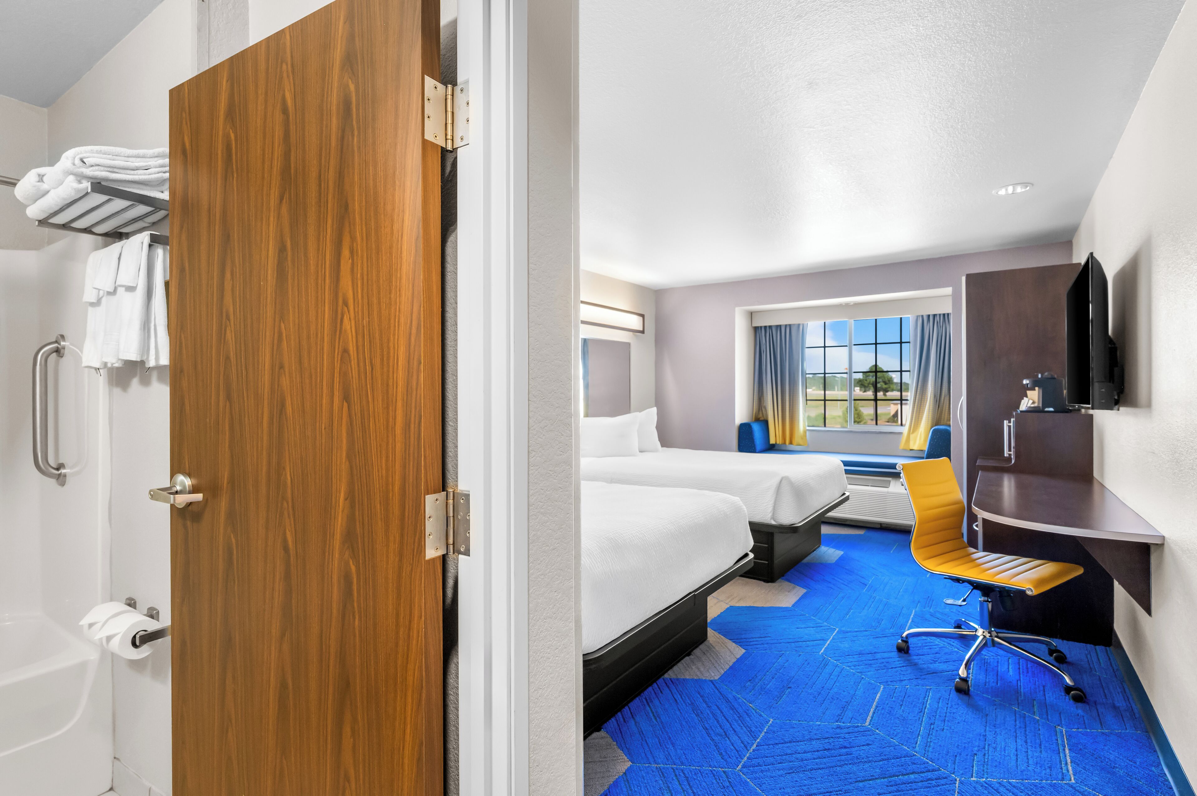 Habitación de hotel con puerta de madera, bañera, dos camas, escritorio, silla y ventana grande con vistas.