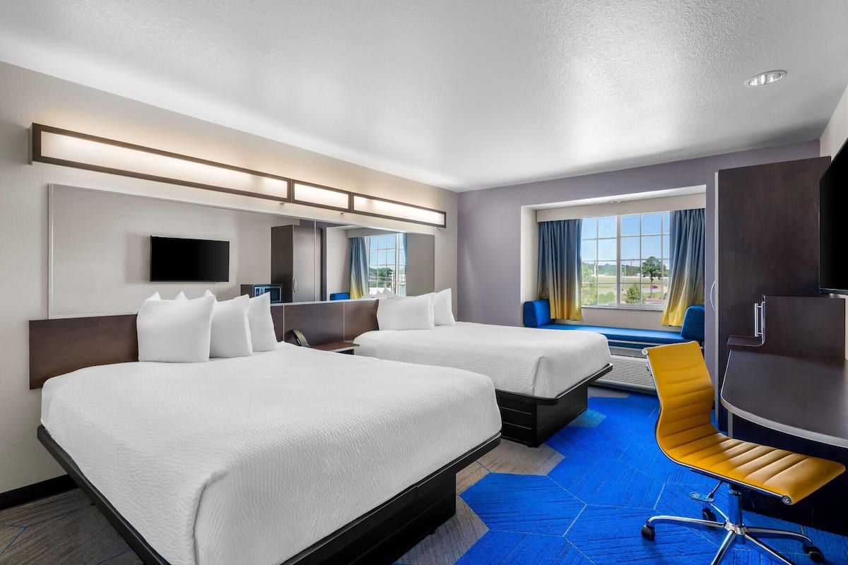 Ein Hotelzimmer mit zwei Betten, einem Schreibtisch, einem Stuhl, einem Fernseher und einem Fenster mit Vorhängen.