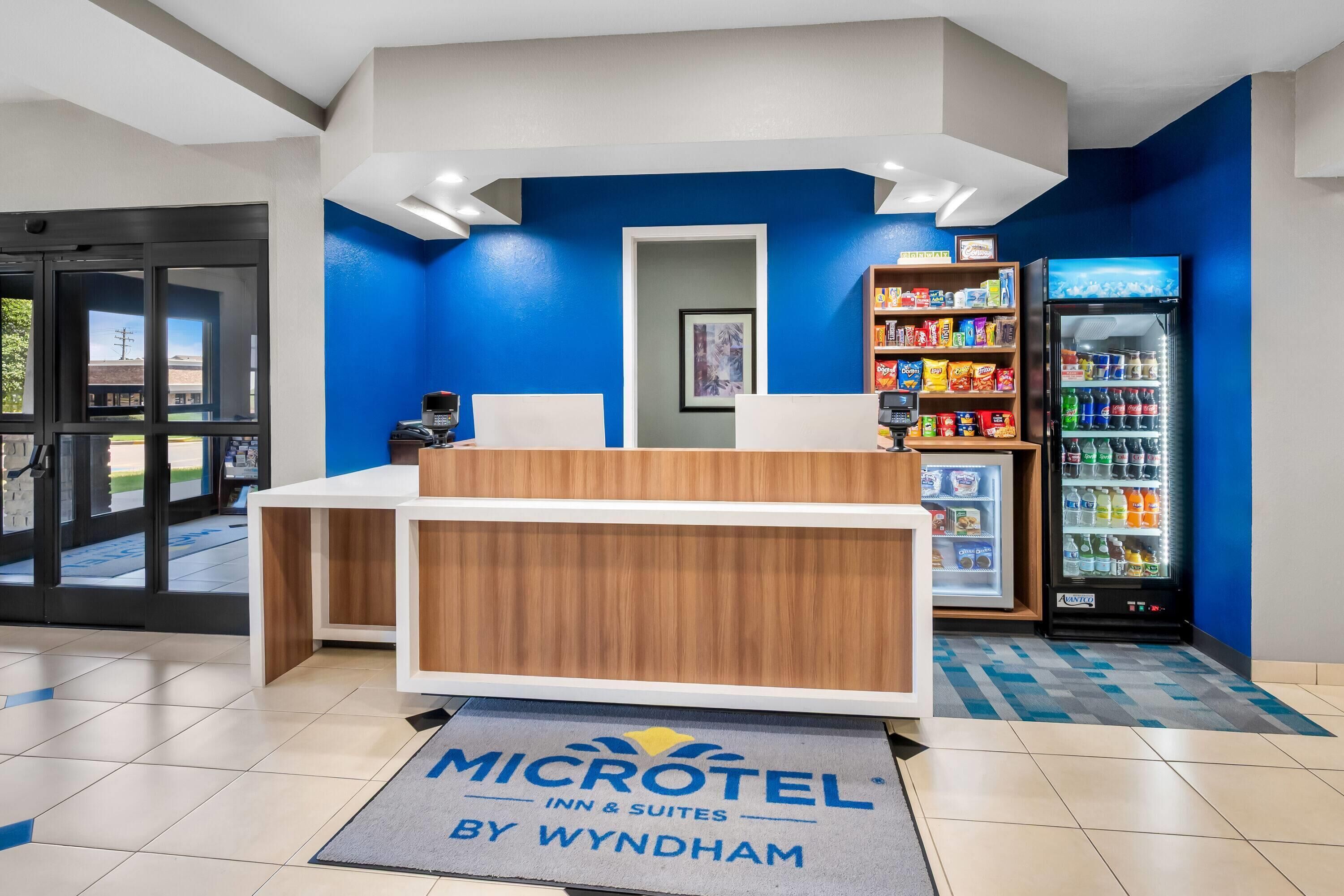 A Microtel Inn & Suites reception area with a blue wall, a vending machine, and a refrigerator stocked with beverages.