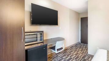 A compact hotel room with a flat-screen TV mounted on the wall, a small kitchenette with a microwave and refrigerator, a foldable desk with a chair, and a patterned carpet.